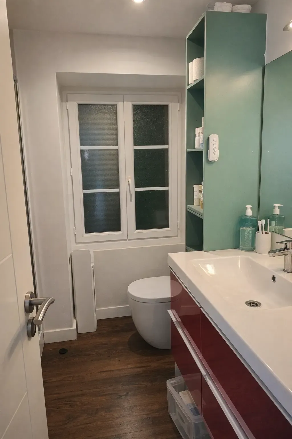 Petite salle de bain rénovée avec meuble bordeaux, fenêtre et étagère verte
